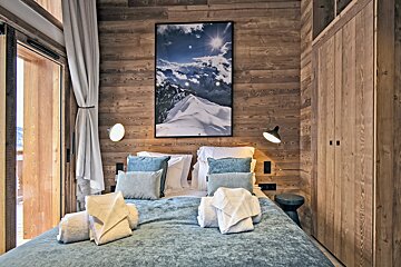 Alpine chalet bedroom with wood-paneled walls, a double bed with blue accents, and a large framed mountain photo.