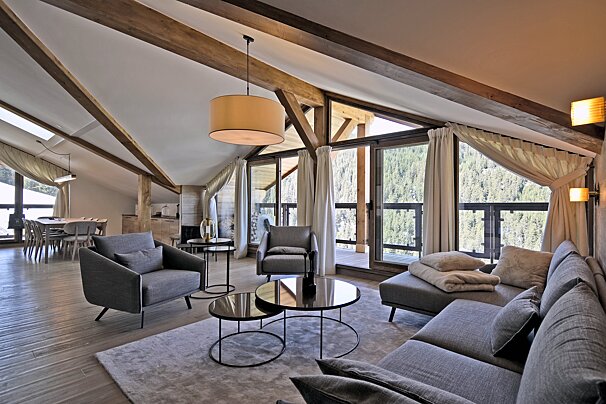 A spacious, modern alpine chalet living room with exposed wood beams, grey furniture, and panoramic forest views.