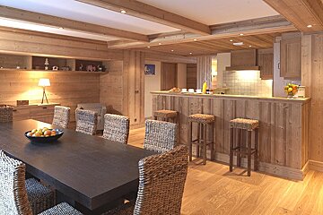 Cozy, rustic wooden chalet-style dining room & kitchen with a long table, wicker chairs, and breakfast bar with stools.