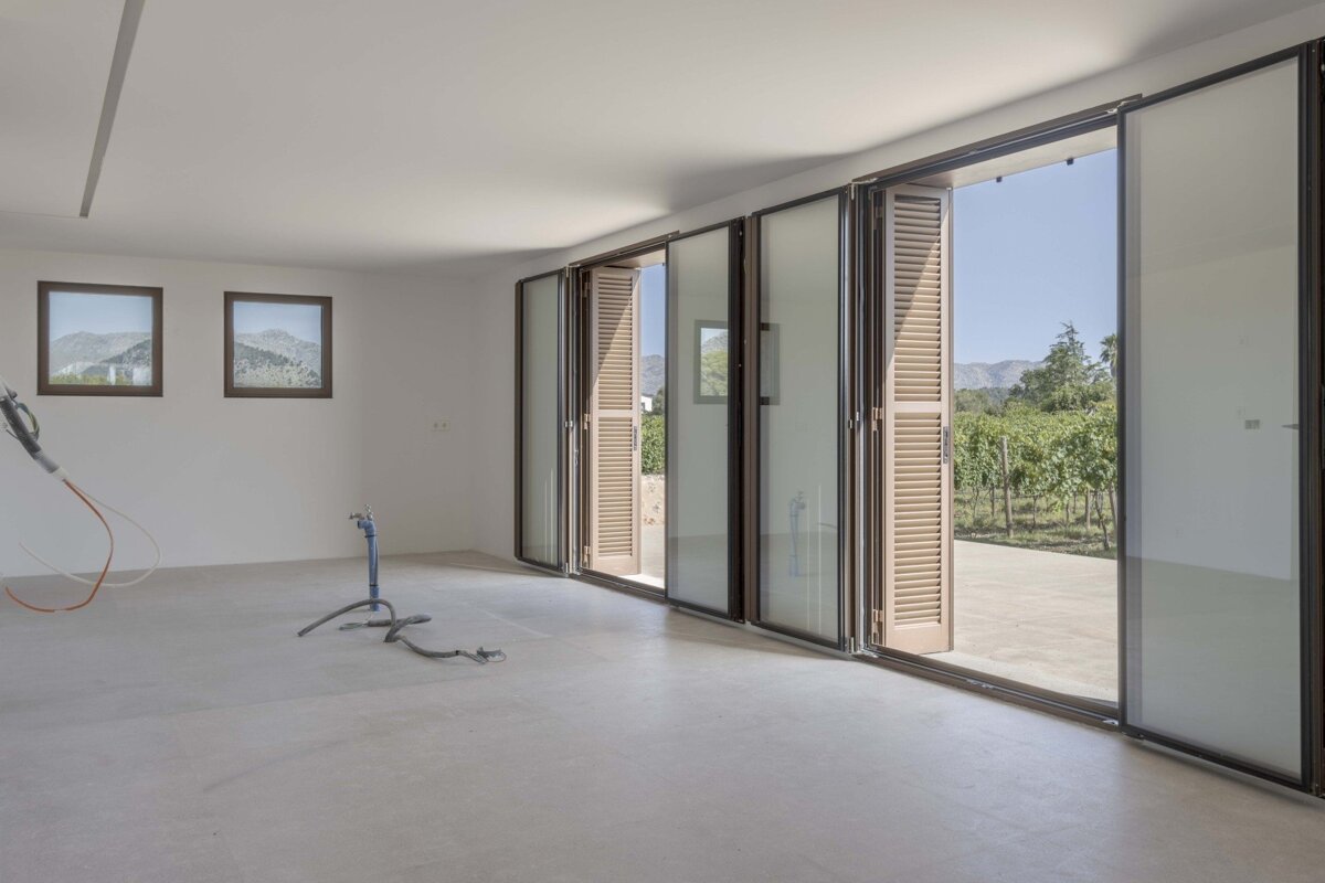Bright, empty room featuring large glass bi-fold doors that open to a vineyard & mountains. Two smaller windows show mountain views, construction items on floor.