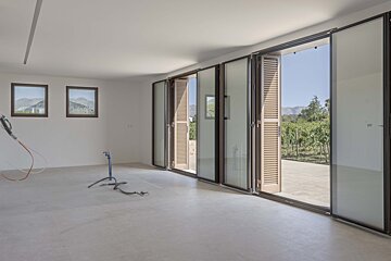 Bright, empty room featuring large glass bi-fold doors that open to a vineyard & mountains. Two smaller windows show mountain views, construction items on floor.