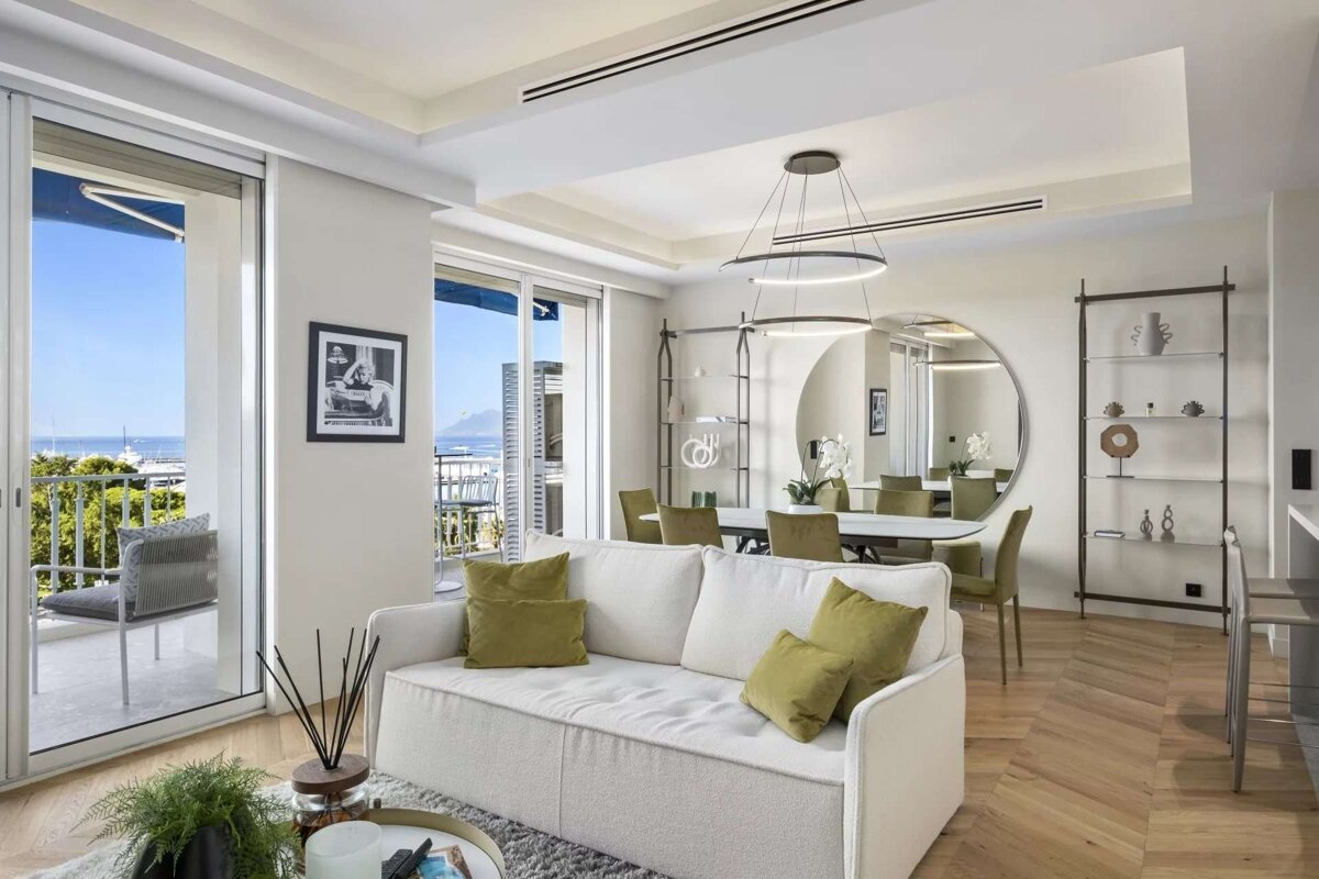 A bright, modern living and dining room features a white sofa, green chairs, stylish decor, and a balcony overlooking the sea.