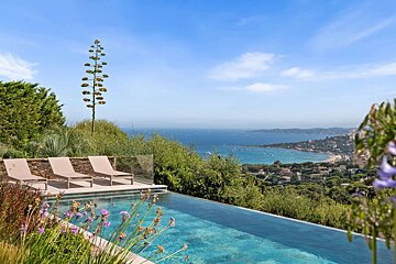 A beautiful infinity pool with sun loungers offers stunning views of a sparkling blue bay, distant coastline, and lush green landscape.