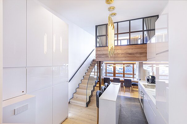Modern loft apartment featuring a sleek white kitchen, wooden stairs leading to a mezzanine, and golden pendant lighting.