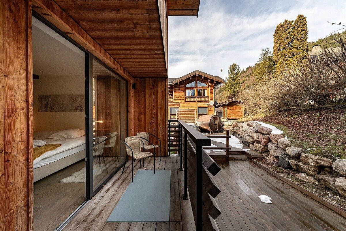 A cozy wooden balcony with glass doors to a bedroom, overlooking a rustic chalet, barrel sauna, and a snowy, wooded mountain landscape.