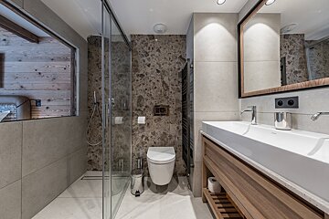 A modern bathroom with a glass shower, toilet, and wood vanity. Features marble-like and grey tiles, plus a peek into a wood-paneled room.