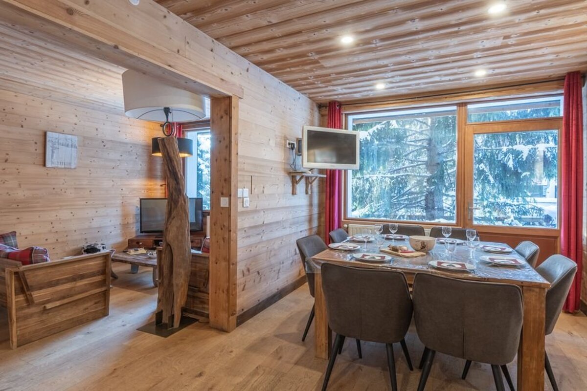 Cozy wooden cabin interior featuring a set dining table, large windows with snowy forest views, and an adjacent living room.