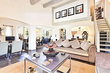A bright, spacious living room with a brown sofa, coffee table, and orange rug. Archways lead to a dining area, and a staircase ascends upstairs.