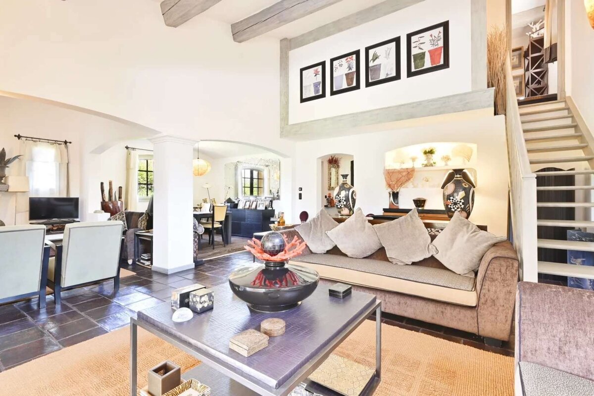 A spacious, open-plan living room featuring a brown sofa, coffee table, and visible dining area. It has high ceilings, exposed beams, a staircase, and various decor pieces.