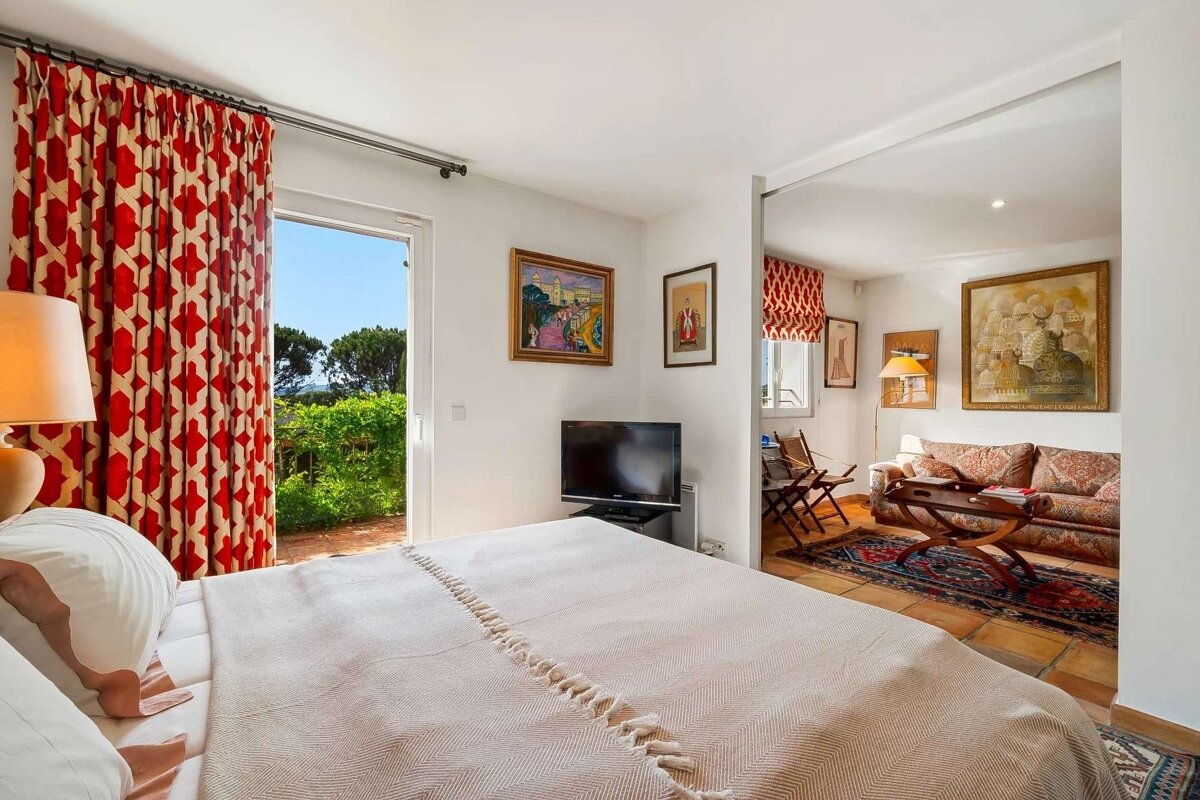 A bright bedroom with a large bed, red patterned curtains opening to a garden, and a TV, connecting to a cozy living area with a couch and art.