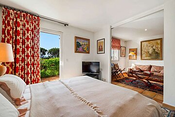 A bright bedroom with a large bed, red patterned curtains opening to a garden, and a TV, connecting to a cozy living area with a couch and art.