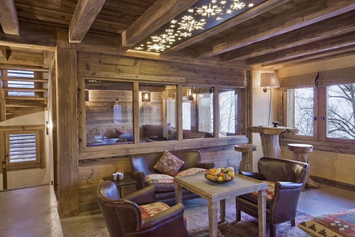 A rustic, cozy chalet living room boasts exposed wooden beams, leather furniture, and a unique snowflake-patterned ceiling light, overlooking winter trees.