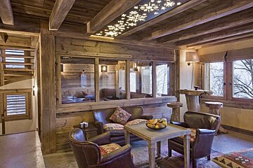 A rustic, cozy chalet living room boasts exposed wooden beams, leather furniture, and a unique snowflake-patterned ceiling light, overlooking winter trees.