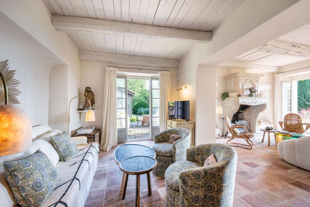 A cozy, rustic living room featuring a beamed ceiling, terracotta floor, eclectic furnishings, a large fireplace, and double doors opening to a lush garden.