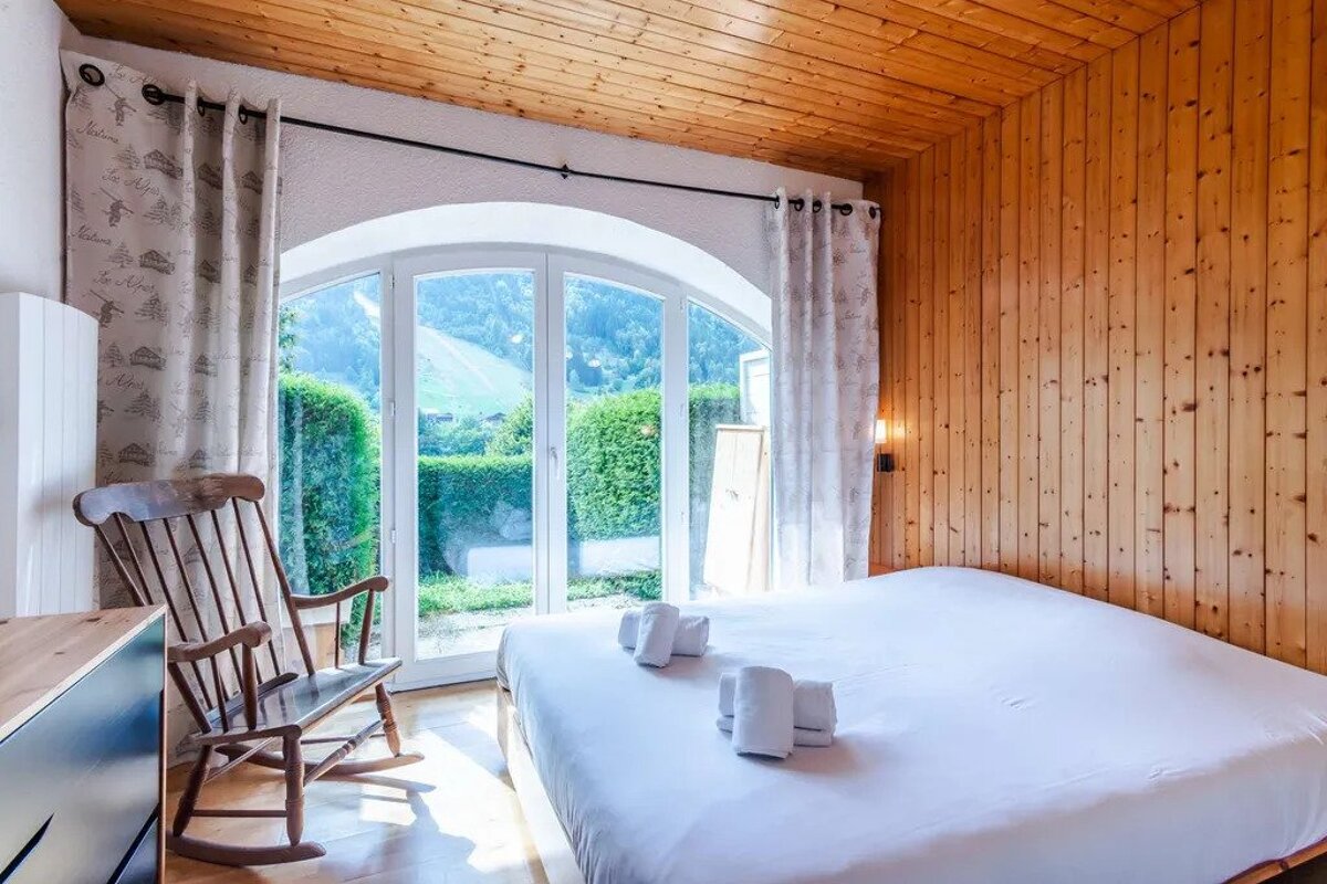 Cozy bedroom with wood-paneled walls, an arched window revealing green hills, a white bed with towels, and a wooden rocking chair.