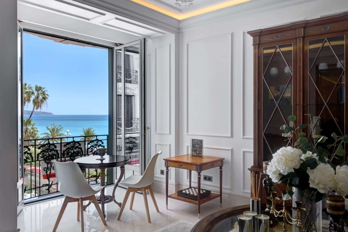 Elegant room with a balcony offering stunning blue ocean views, palm trees, and a coastal town. Features classic white interior design.