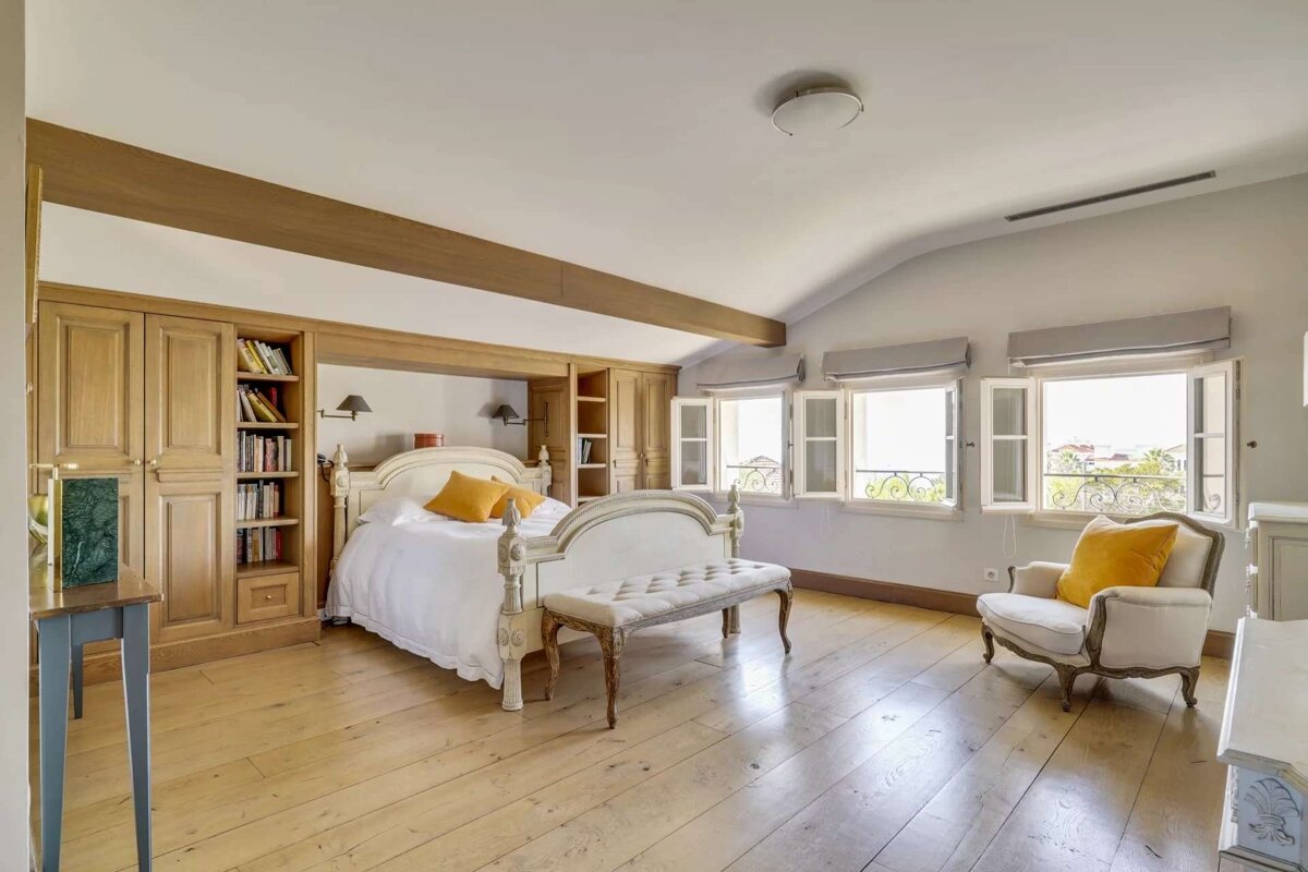 Bright, airy bedroom featuring a white bed with yellow accents, light wood floors, built-in shelving, multiple windows with open shutters, and classic white furniture.