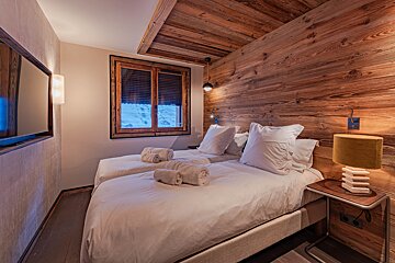 Cozy cabin bedroom featuring twin beds, warm wood-paneled walls and ceiling, and a window looking out onto a snowy landscape.