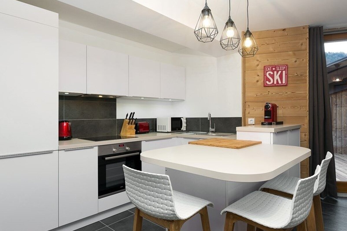 A modern white kitchen featuring an island with woven stools, wire pendant lights, and a wood accent wall with an "EAT SLEEP SKI" sign.