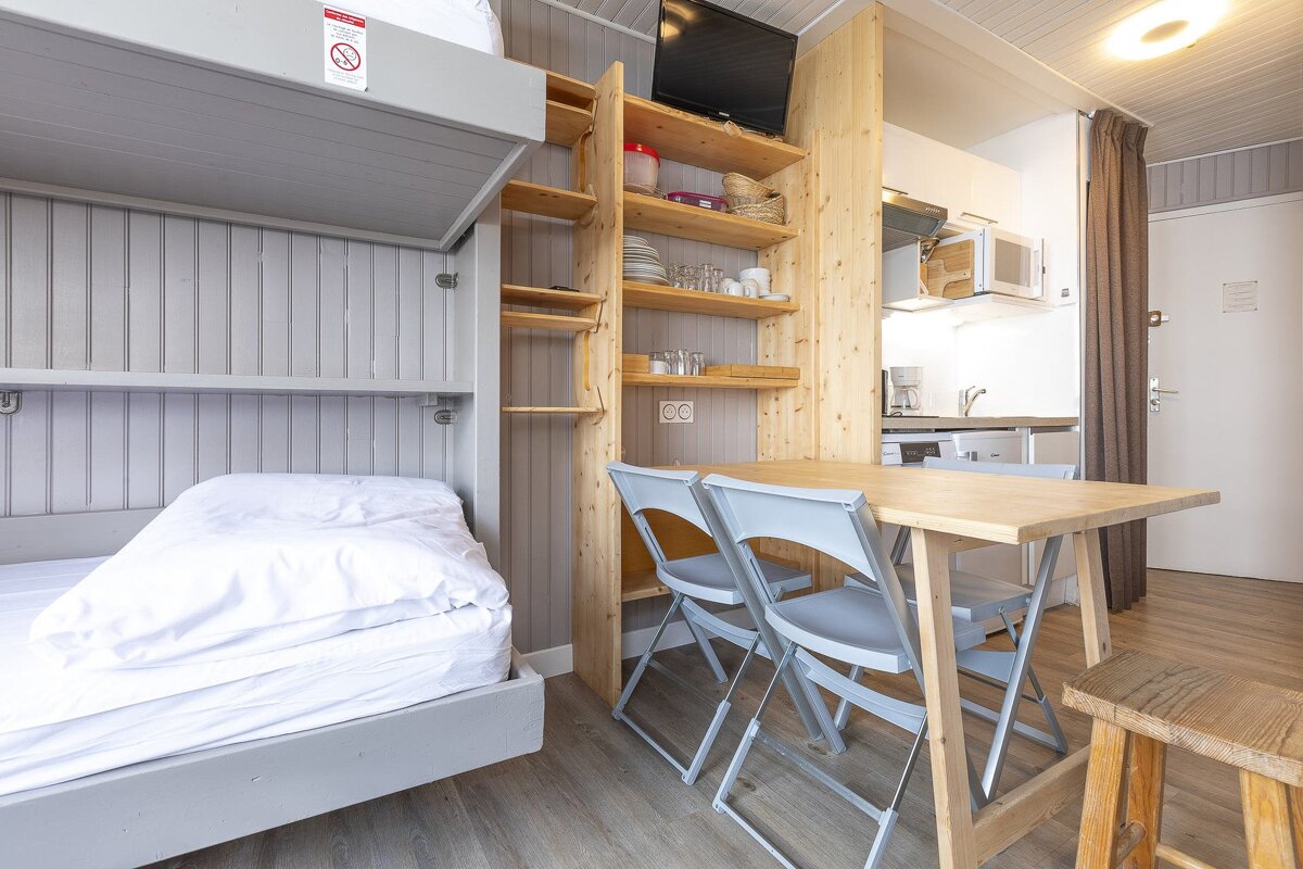 Compact studio with a bunk bed, wooden shelves, dining table, and kitchenette. Light grey walls and natural wood create a functional, cozy space.