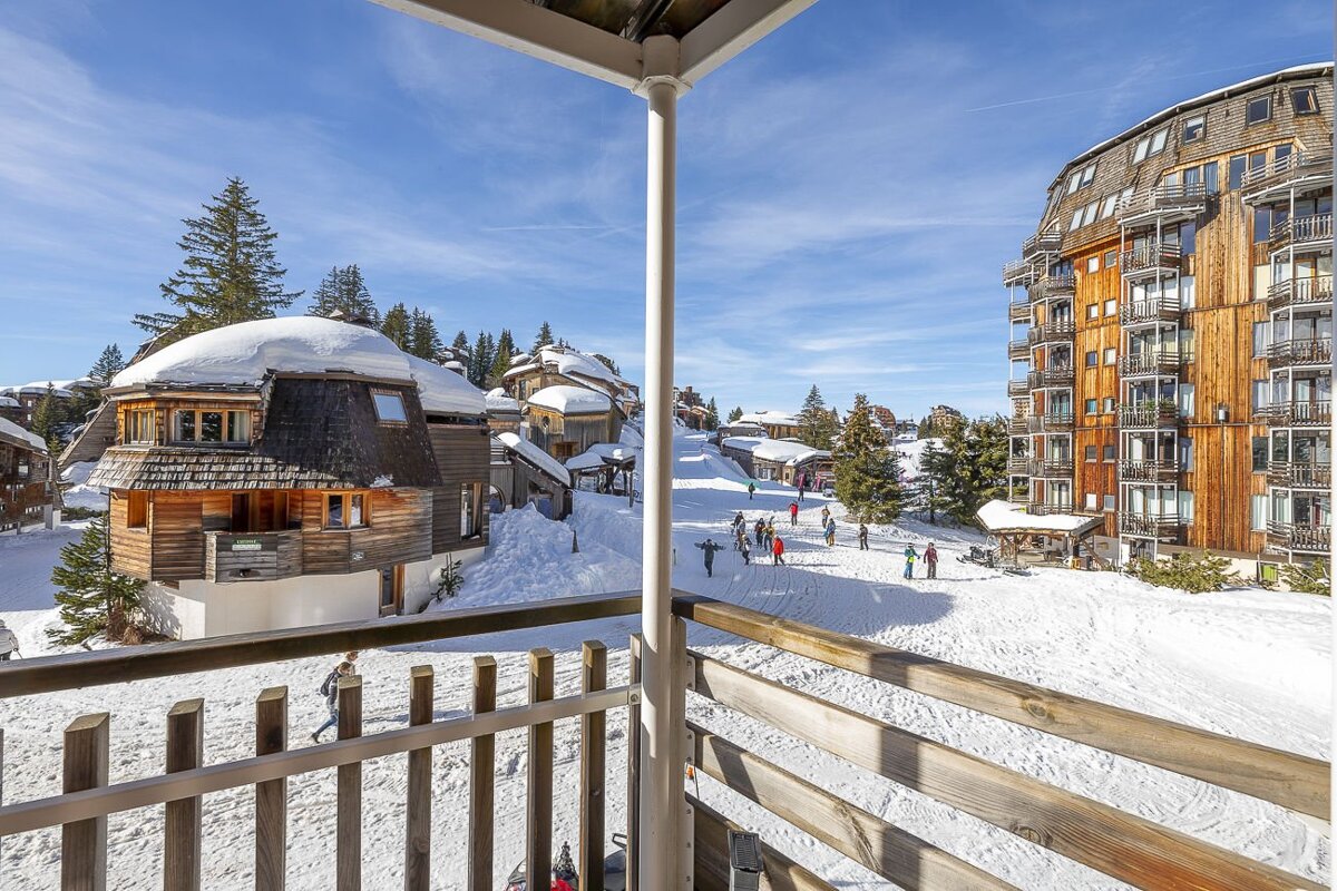 Sunny balcony view of a snowy ski village with wooden chalets, pine trees, and people enjoying the winter day under a blue sky.