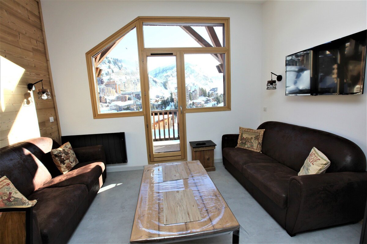 Cozy living room with two brown sofas, coffee table, and a large window offering a snowy mountain village view. A TV is mounted on the wall.