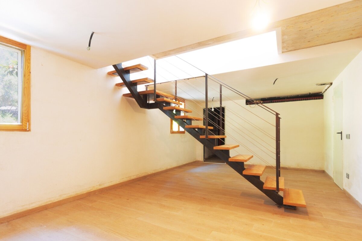 A modern room with light wood flooring, white walls, and a stylish floating staircase with wooden treads and metal railing. A bright window is on the left.