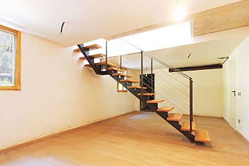 A modern room with light wood flooring, white walls, and a stylish floating staircase with wooden treads and metal railing. A bright window is on the left.