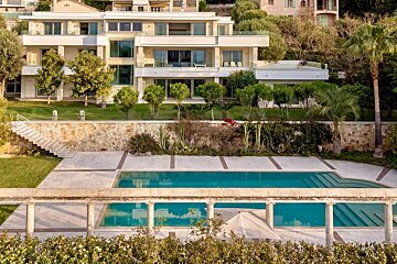 Elevated view of a luxurious modern white villa with balconies, a large blue swimming pool, terraced gardens, green lawns, and diverse trees.