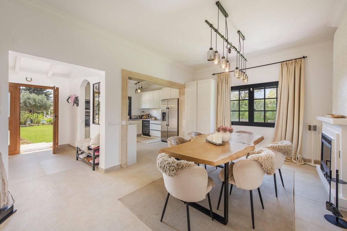 An inviting open-plan home with a rustic dining table, plush chairs, modern kitchen, and an entryway to a green garden.