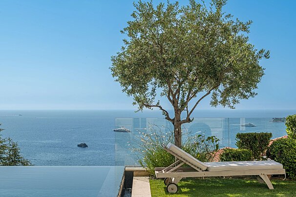 A lounge chair under an olive tree next to an infinity pool with a stunning ocean view.