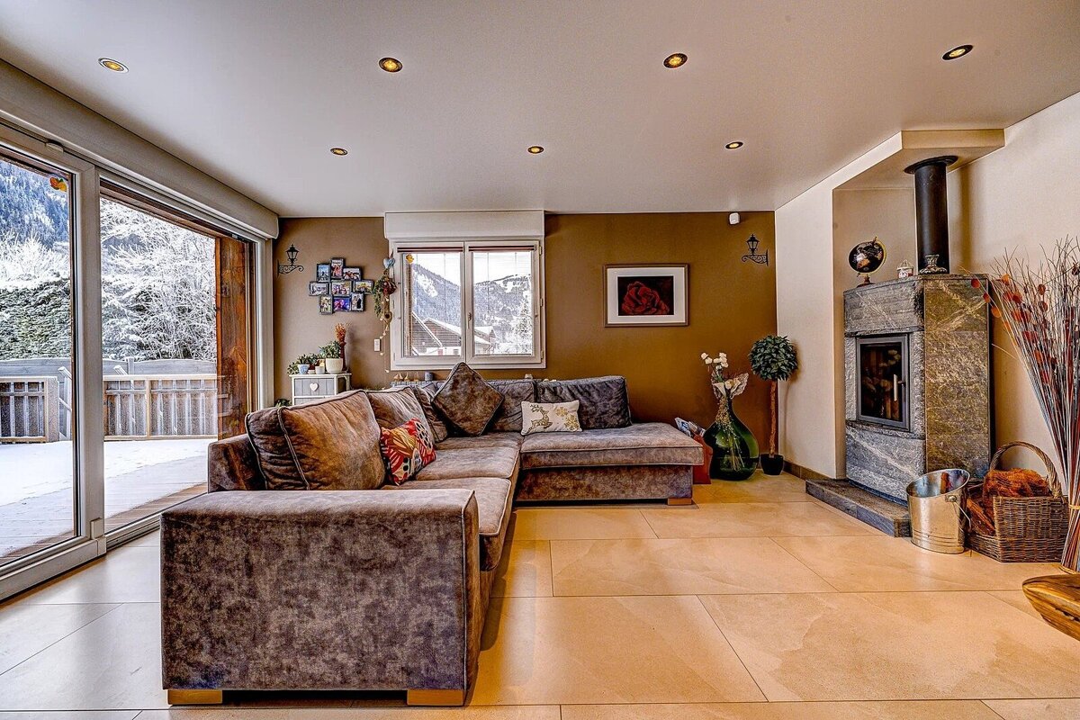 A cozy living room with a large sofa, stone fireplace, and a snowy mountain view through big glass doors, offering warmth amidst winter.