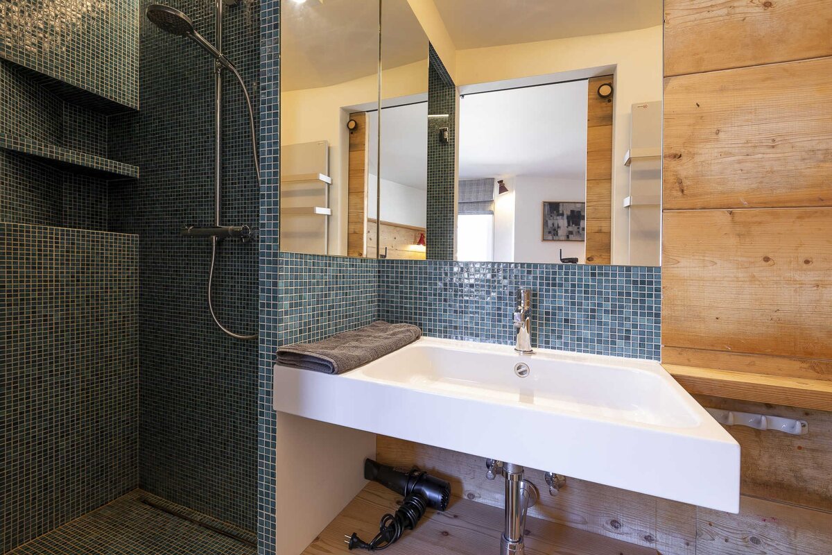 Modern bathroom featuring a white sink, teal mosaic tiled shower, large mirror, and wooden wall panels.