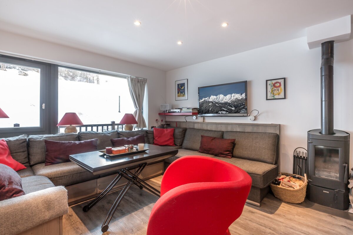 A cozy living room with an L-shaped sofa, red chair, fireplace, and a large window offering snowy mountain views. A TV displays a mountain scene.