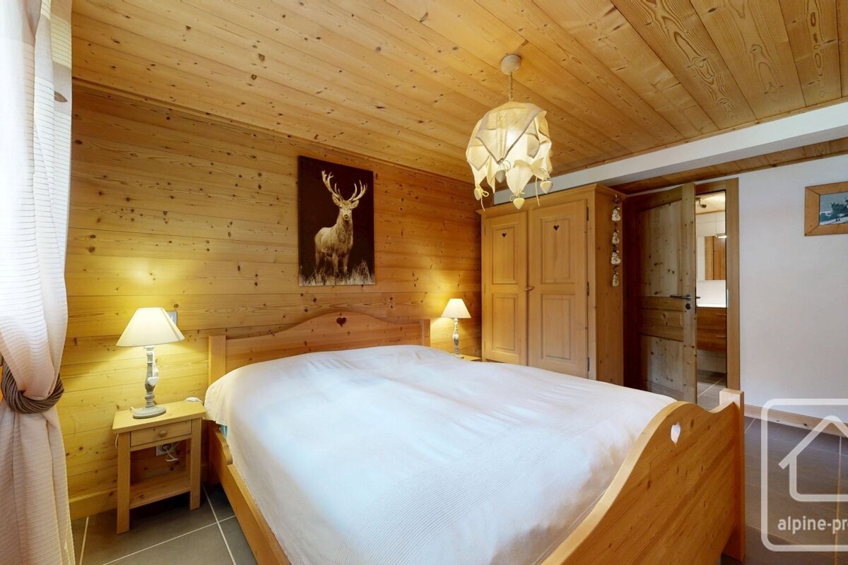 Rustic wood-paneled bedroom with a large bed, deer art, wooden wardrobe, and bedside lamps. Bathroom door open.