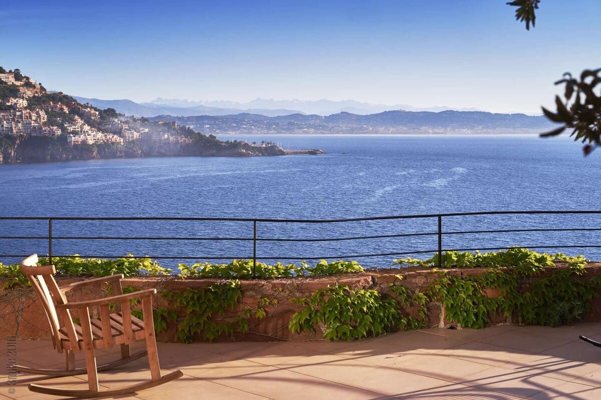 A sunlit patio with a wooden rocking chair overlooks a vivid blue sea, a charming hillside town, and misty mountains in the distance.