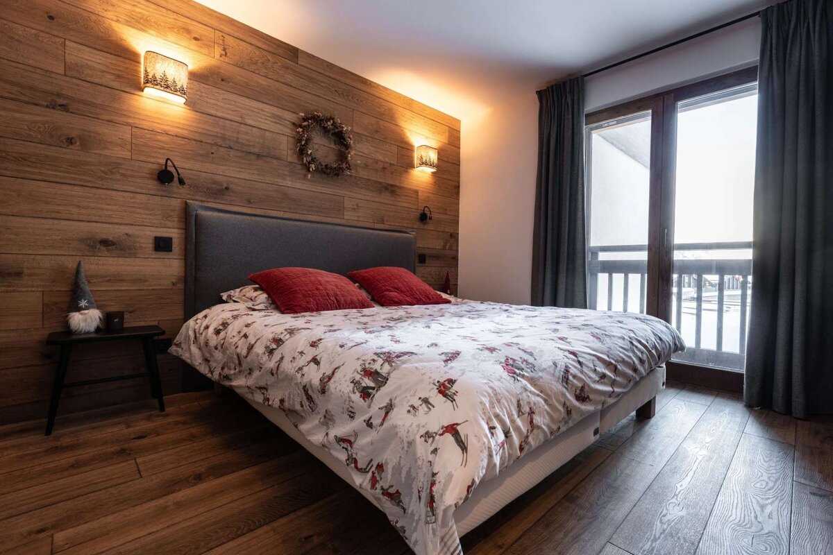 A warm, inviting bedroom featuring wood paneling, a ski-themed duvet with red pillows, and a large window looking out, evoking a chalet feel.