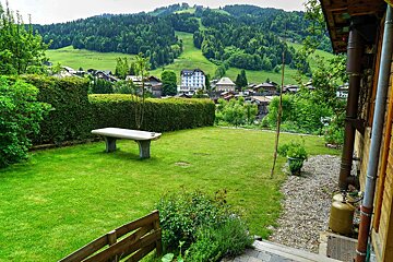 A lush green garden with a stone bench and hedge, framing a village set against forested mountains and a ski slope.