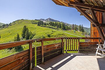 Sunny mountain view from a wooden balcony, showcasing green slopes, pine trees, ski lifts, and snowy peaks under a clear sky.