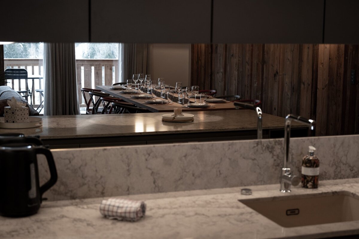 A modern kitchen counter and sink, with a mirrored backsplash reflecting a long wooden dining table set for a meal.