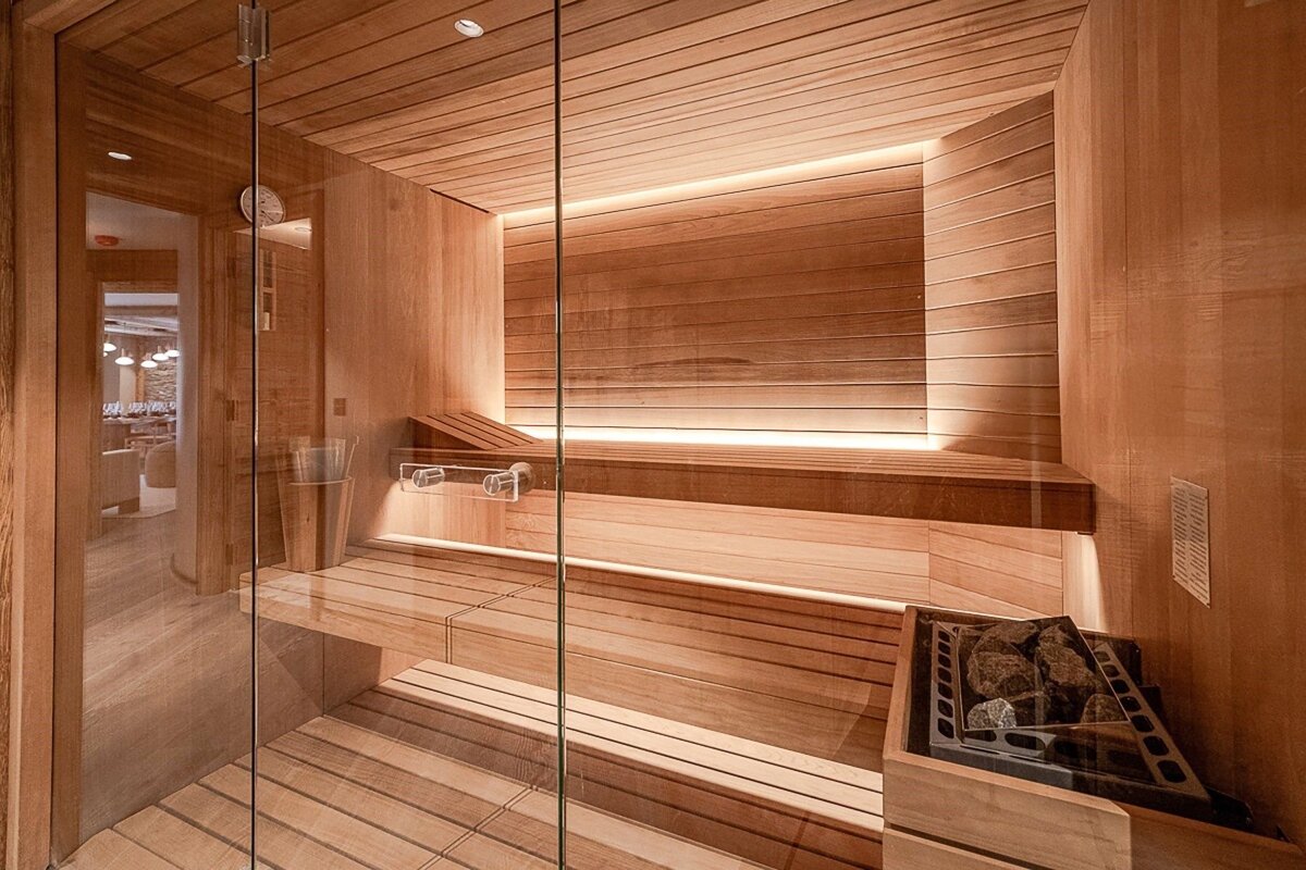 A modern, warm-toned wooden sauna with glass doors, multi-level benches, soft backlighting, and a heater with sauna stones.