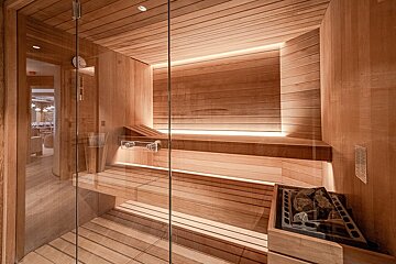 A modern, warm-toned wooden sauna with glass doors, multi-level benches, soft backlighting, and a heater with sauna stones.