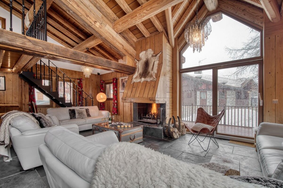 Cozy wooden chalet living room with a warm fireplace, white sofas, and a large window framing a snowy outdoor scene.