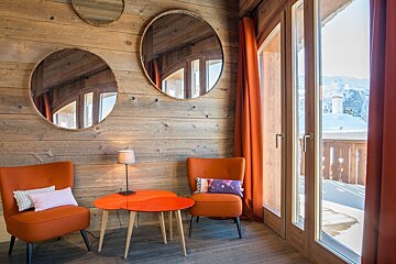 Rustic wood-paneled room with orange chairs, a table, and mirrors, offering a snowy mountain view from a balcony with bright orange curtains.