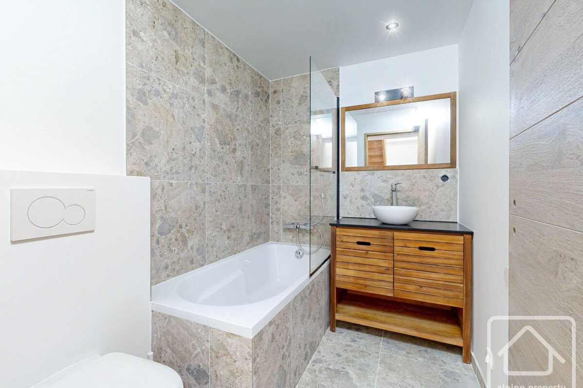 A modern bathroom featuring a bathtub with glass screen, wooden vanity with vessel sink, toilet, and beige marble-look tiles.