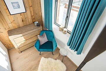 A cozy room with wooden walls, a teal armchair, and matching curtains. Sunlight streams onto the light wood floor, illuminating a white rug and a window view of snow.