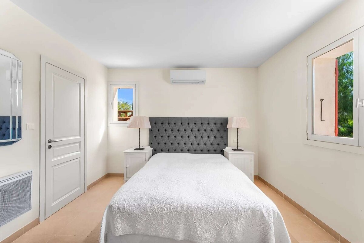 A bright, clean bedroom features a bed with a grey tufted headboard, white linens, two lamps on nightstands, an AC unit, and windows.