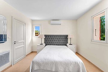 A bright, clean bedroom features a bed with a grey tufted headboard, white linens, two lamps on nightstands, an AC unit, and windows.