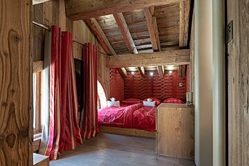 A cozy, rustic bedroom featuring a bed with red bedding and a patterned red headboard, exposed wooden beams, and red striped curtains.