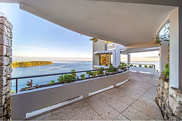 A curved outdoor walkway/balcony with planters, offering a panoramic view of a serene blue ocean and distant land under a clear sky.